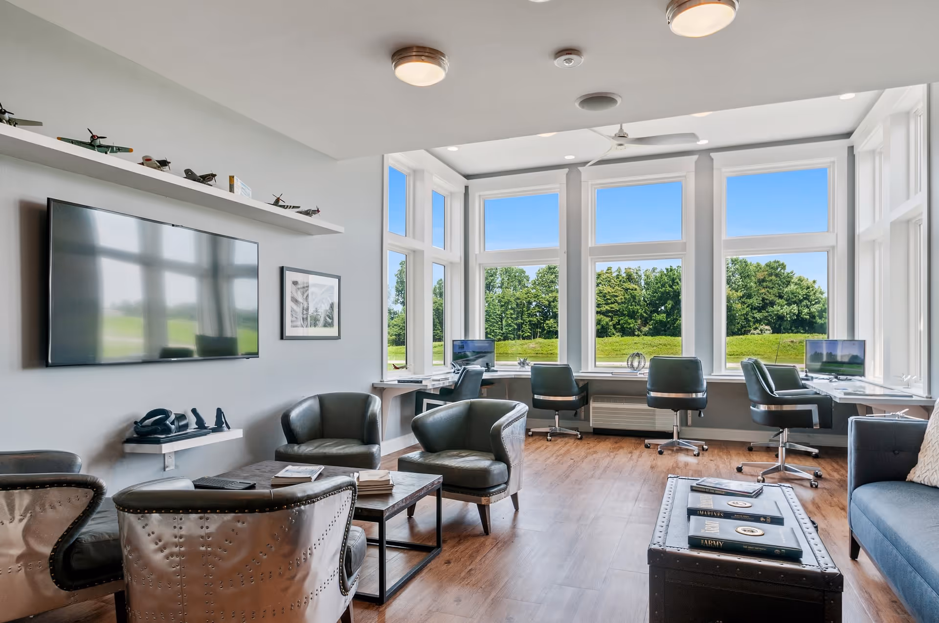 A bright and modern common area with large windows showing a green outdoor view. The room features several leather chairs arranged around a coffee table, a wall-mounted flat-screen TV, and a shelf with model airplanes. There are also desks with computers positioned by the windows, and the floor is wooden.