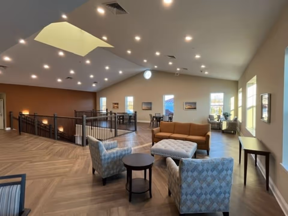 A spacious, well-lit common area with a high ceiling featuring recessed lighting and a skylight. The room has a brown accent wall and several windows letting in natural light. There is a seating arrangement with a brown sofa, two patterned armchairs, and a large ottoman in the center. In the background, there are tables and chairs near the windows, and a railing around a stairwell.