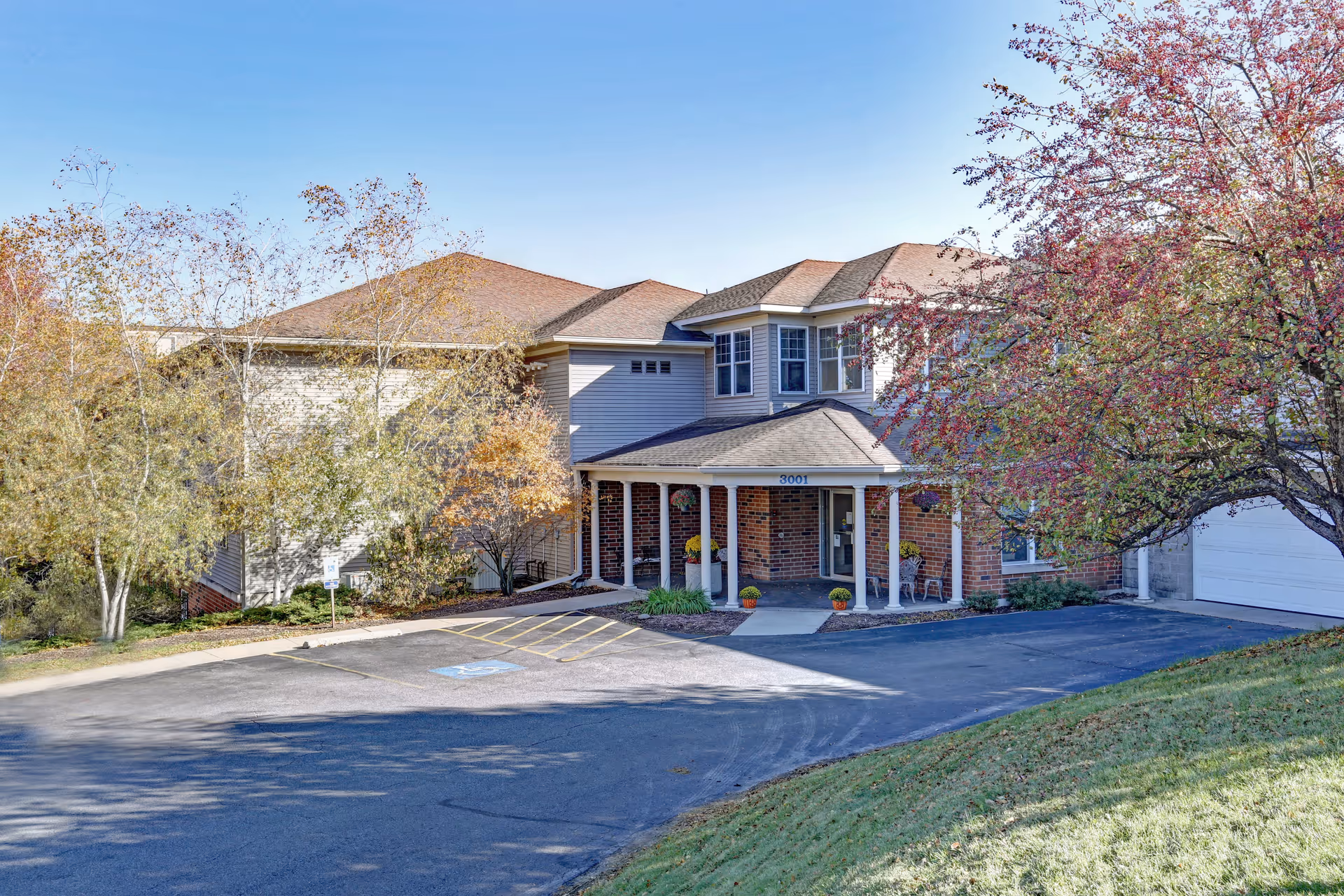 Exterior view of a two-story senior living facility building with a covered entrance, surrounded by trees with autumn foliage and a parking area with a handicapped parking space.