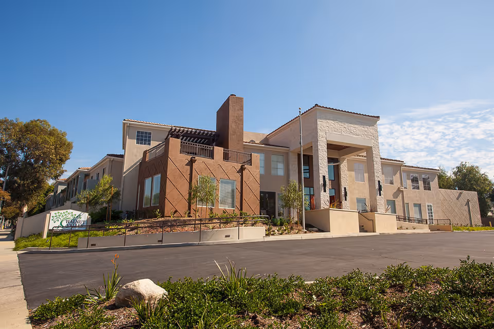 Exterior view of Walnut Valley Senior Living facility, showing a large two-story building with beige and brown walls, multiple windows, and a prominent entrance with a covered porch. The building is surrounded by landscaped greenery and a paved driveway under a clear blue sky.