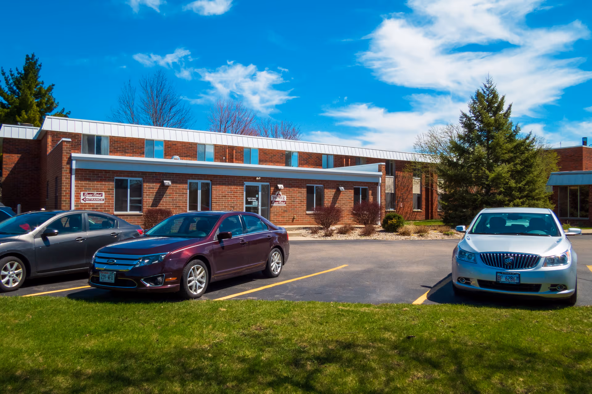 Exterior view of a brick senior living facility building with several windows and a white roof trim under a blue sky with scattered clouds. There are three cars parked in front of the building on an asphalt parking lot, with a green grassy area in the foreground. Trees and shrubs are visible around the building.