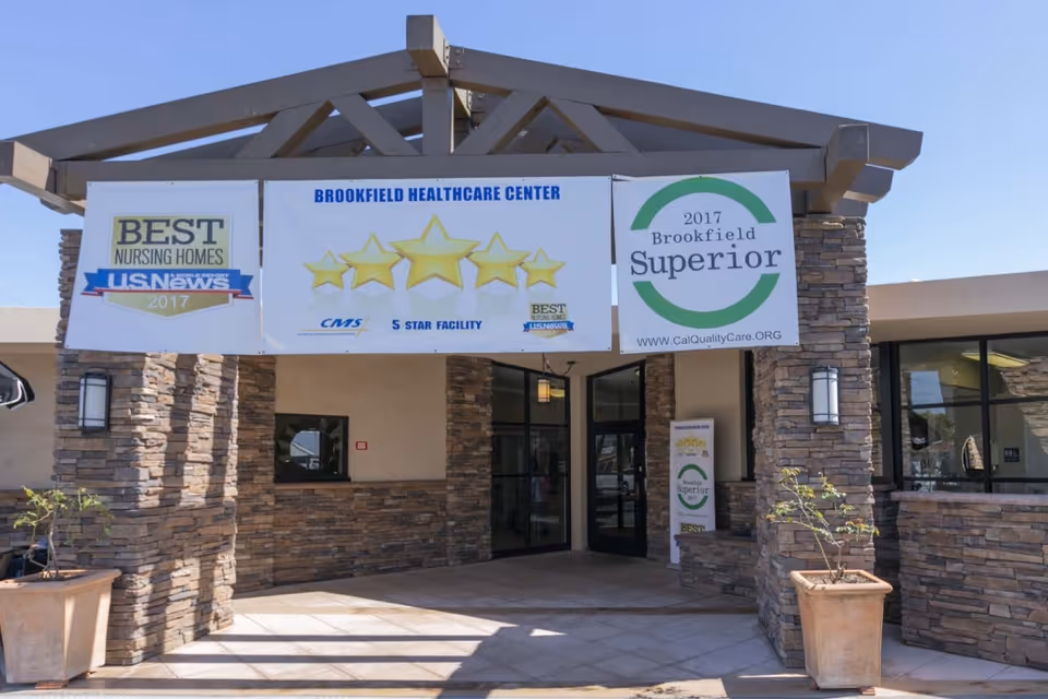 Entrance of Brookfield Healthcare Center with stone pillars and a covered walkway. Banners hang above the entrance displaying awards including 'Best Nursing Homes US News 2017', a 5-star facility rating, and a '2017 Brookfield Superior' quality care award.