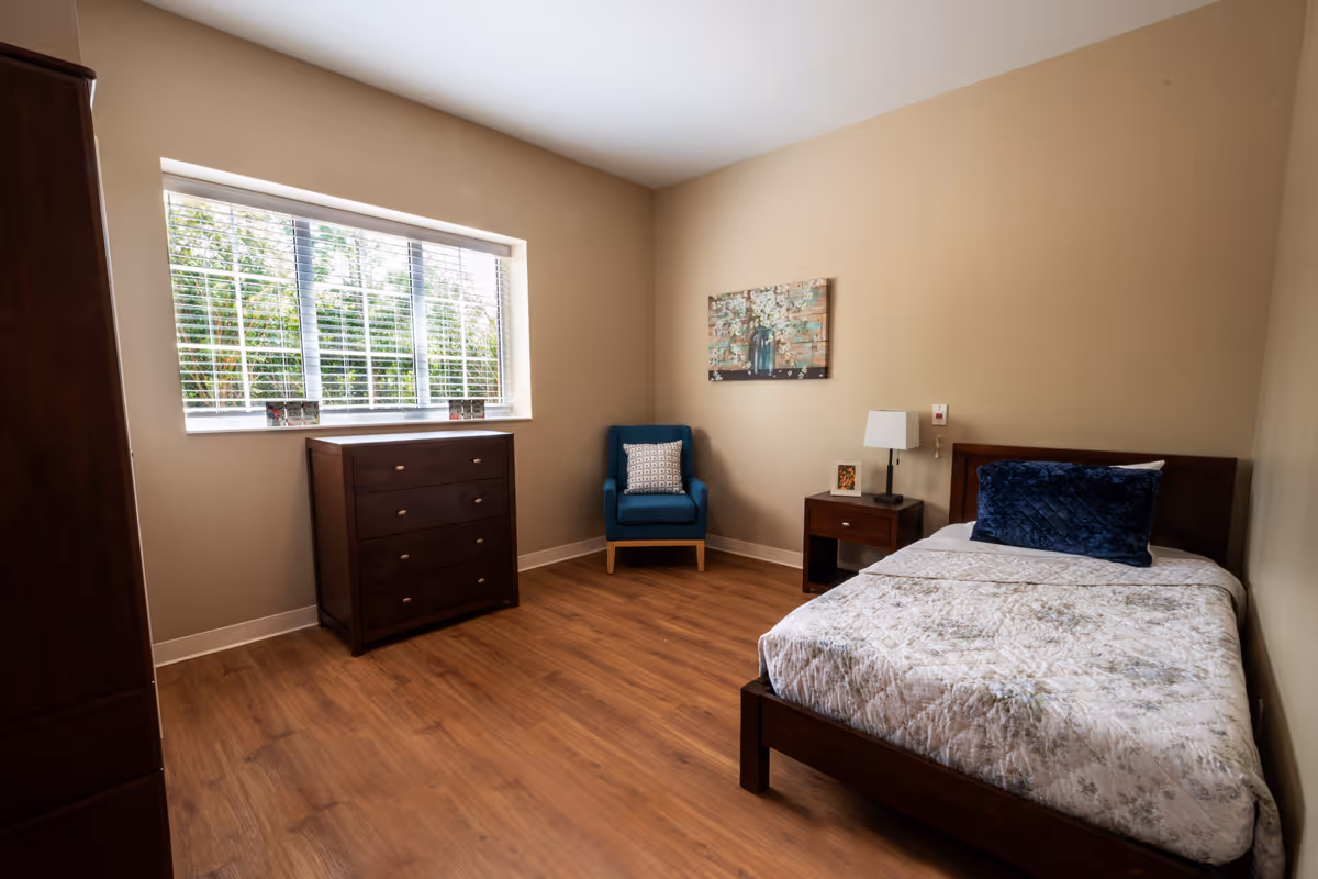 A simple, clean bedroom with a single bed featuring a white and gray patterned comforter and a dark blue pillow. The room has wooden flooring, beige walls, a wooden dresser under a window with white blinds, a blue armchair with a patterned cushion, a wooden nightstand with a lamp and a small framed picture, and a floral painting on the wall.