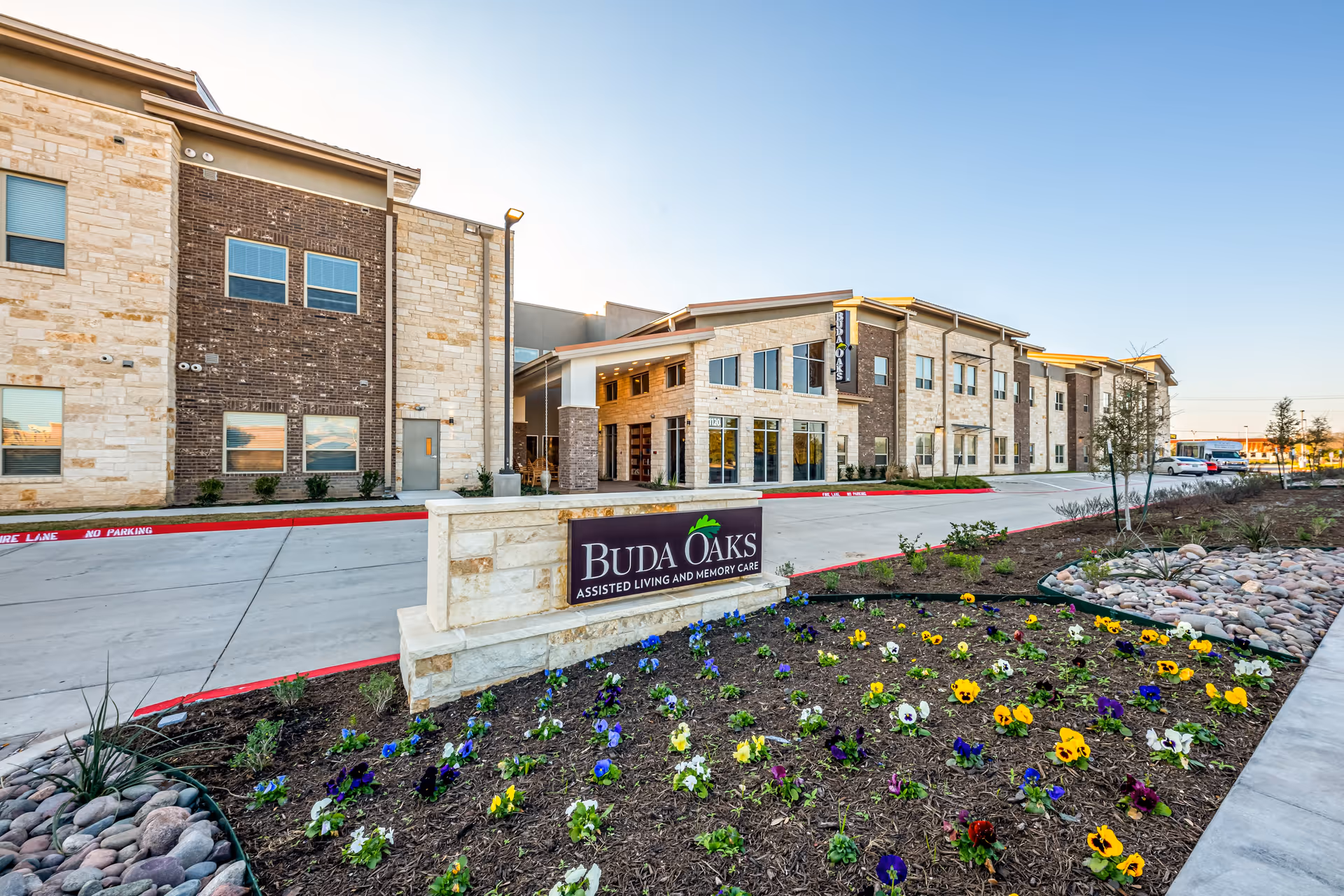 Exterior view of Buda Oaks Assisted Living and Memory Care facility with a stone and brick facade, a driveway, landscaped flower beds with colorful flowers, and clear blue sky.