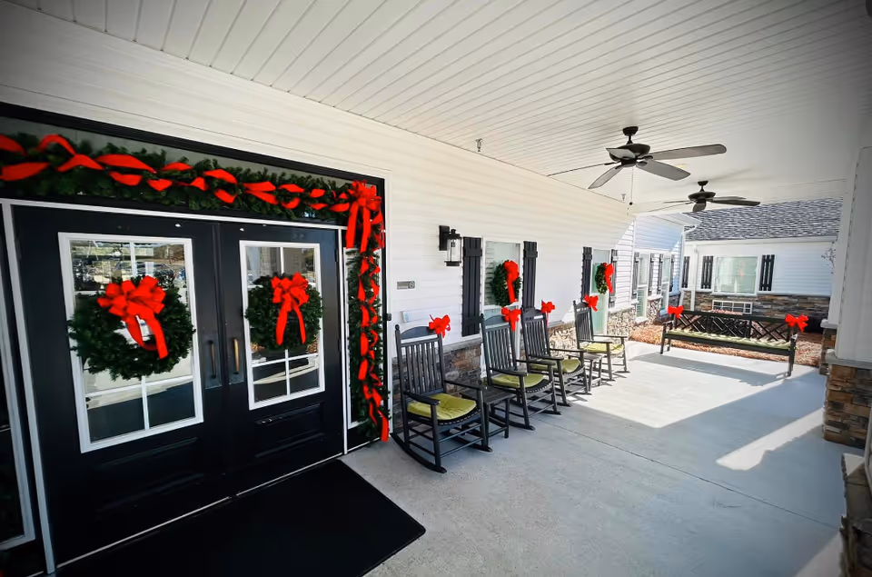 Covered outdoor porch area decorated for the holidays with green wreaths and red bows on black double doors, windows, rocking chairs with green cushions, and a bench. The porch has ceiling fans and stone accents on the lower walls.