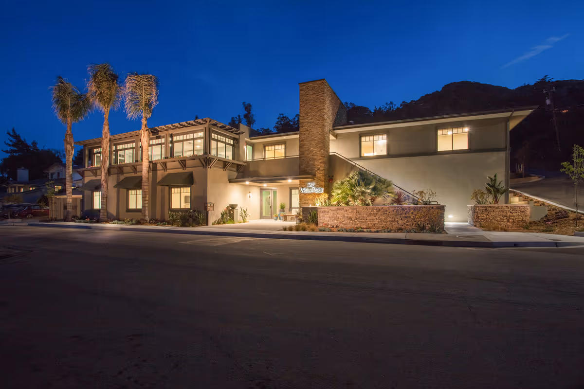 Exterior nighttime view of a two-story building with illuminated windows and entrance, surrounded by palm trees and landscaping, identified as Vista Rosa Elder Care.