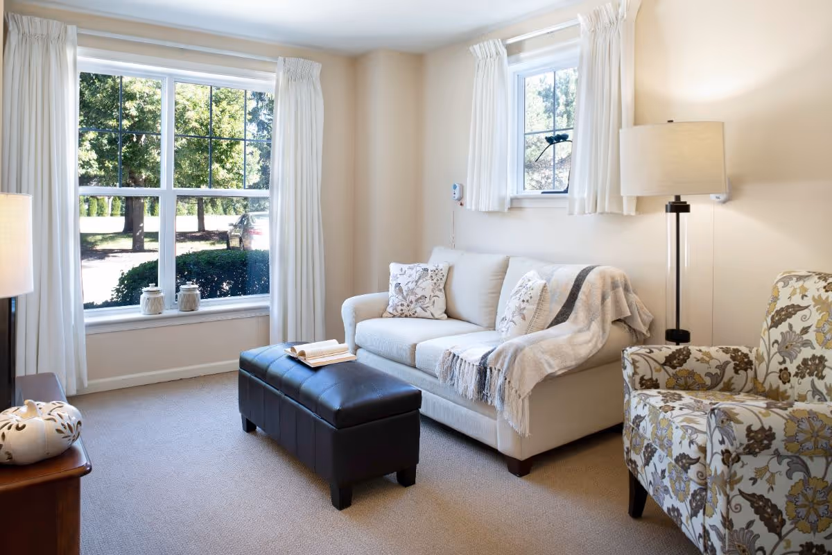 A cozy living room with a beige sofa adorned with patterned cushions and a throw blanket, a floral armchair, a black leather ottoman with an open book on top, a wooden side table with decorative items, two large windows with white curtains letting in natural light, and a floor lamp.