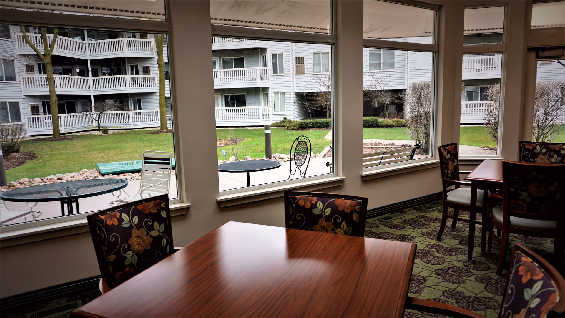 Interior view of a senior living facility dining area with wooden tables and floral upholstered chairs. Large windows provide a view of an outdoor courtyard with green grass, patio tables, chairs, and a multi-story building with balconies.