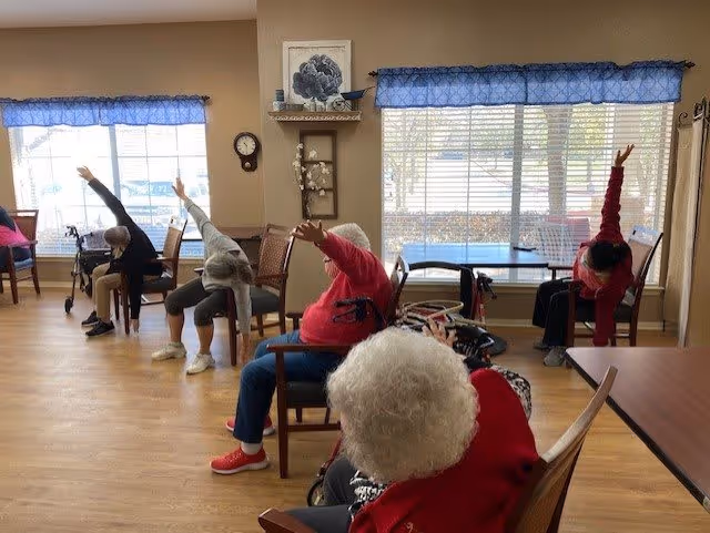 A group of elderly individuals seated in chairs in a spacious room with large windows, participating in a seated stretching exercise with their arms raised and leaning to one side. The room has wooden flooring, beige walls, and blue valance curtains on the windows.