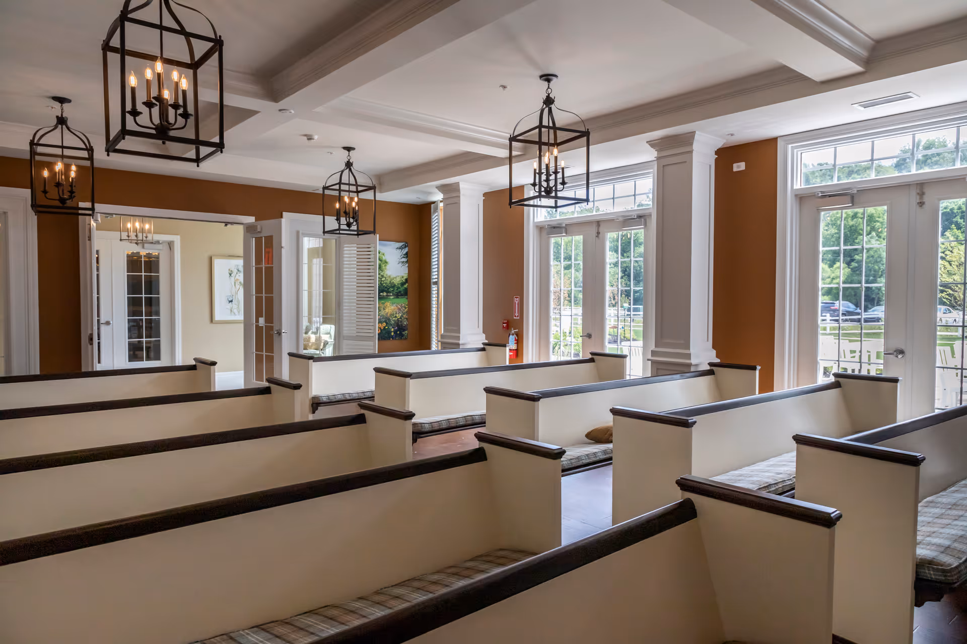Interior view of a room with multiple white wooden pews with plaid cushions, large windows and glass doors letting in natural light, and decorative hanging lantern-style chandeliers. The walls are painted in warm tones, and there are white columns and trim details. Outside greenery is visible through the windows.