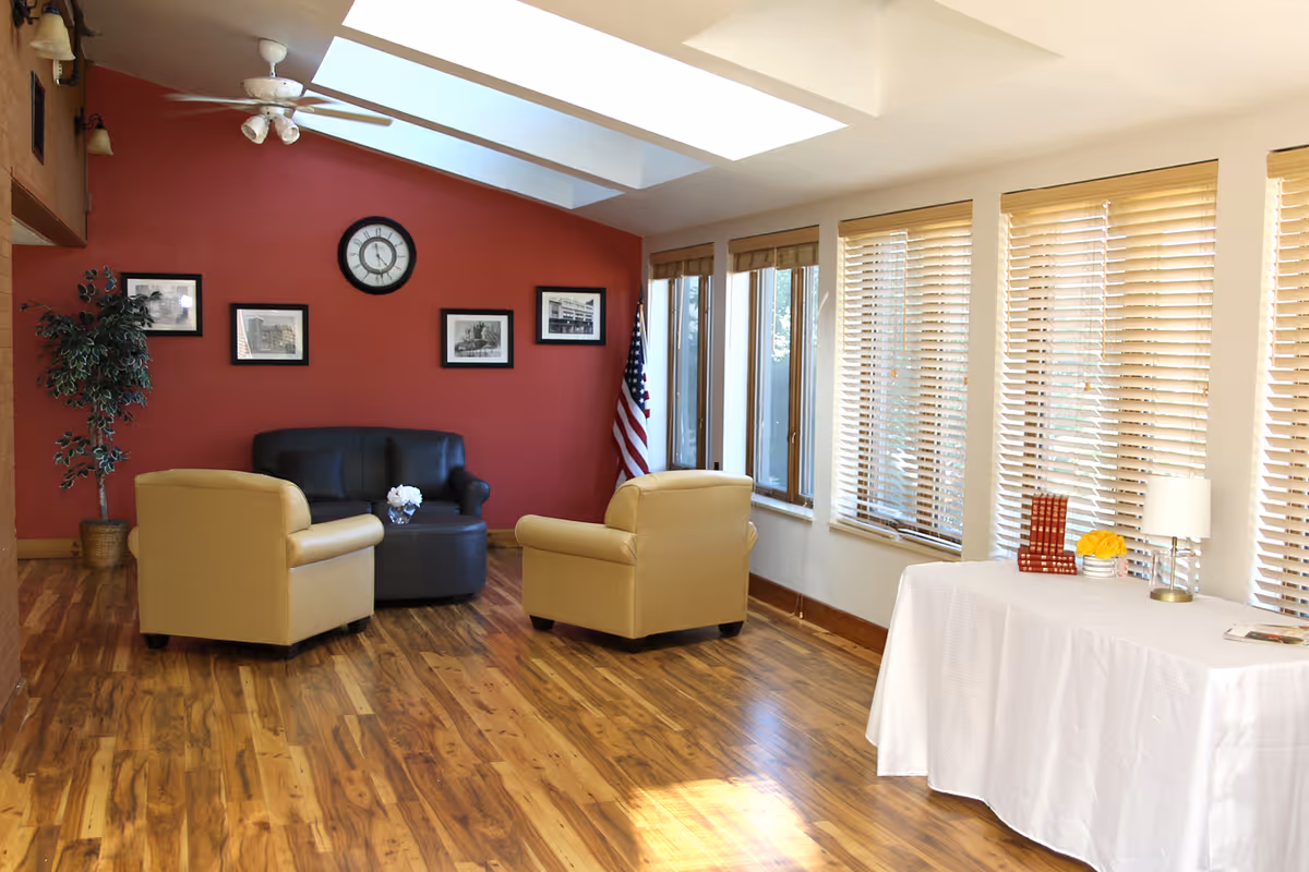 A bright sitting area with wooden flooring, featuring two beige armchairs and a black loveseat arranged around a small round table with a flower vase. The back wall is painted red and decorated with a clock and four framed pictures. There is an American flag in the corner and a potted plant to the left. On the right side, large windows with wooden blinds let in natural light, and a table covered with a white cloth holds a lamp, books, and a small flower arrangement.