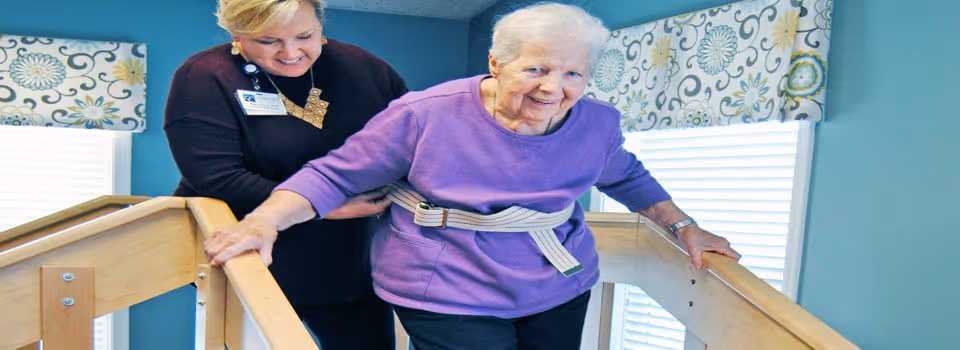 An elderly woman wearing a purple sweater is walking with the assistance of a caregiver who is supporting her from behind. They are using wooden handrails in a well-lit room with blue walls and patterned window valances.