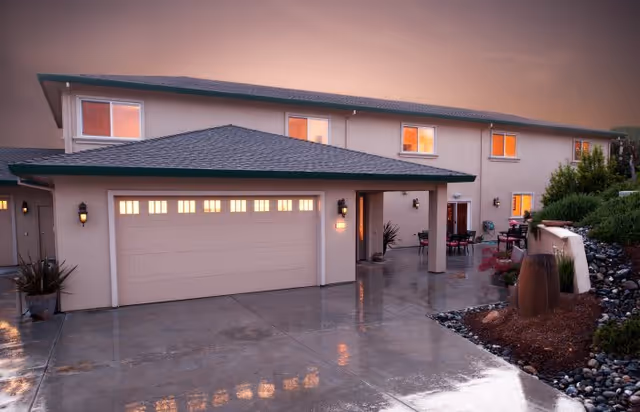 Exterior view of a two-story residential building with a large garage door, illuminated windows, and a wet driveway reflecting the lights. There are plants and landscaping features near the entrance and along the side.