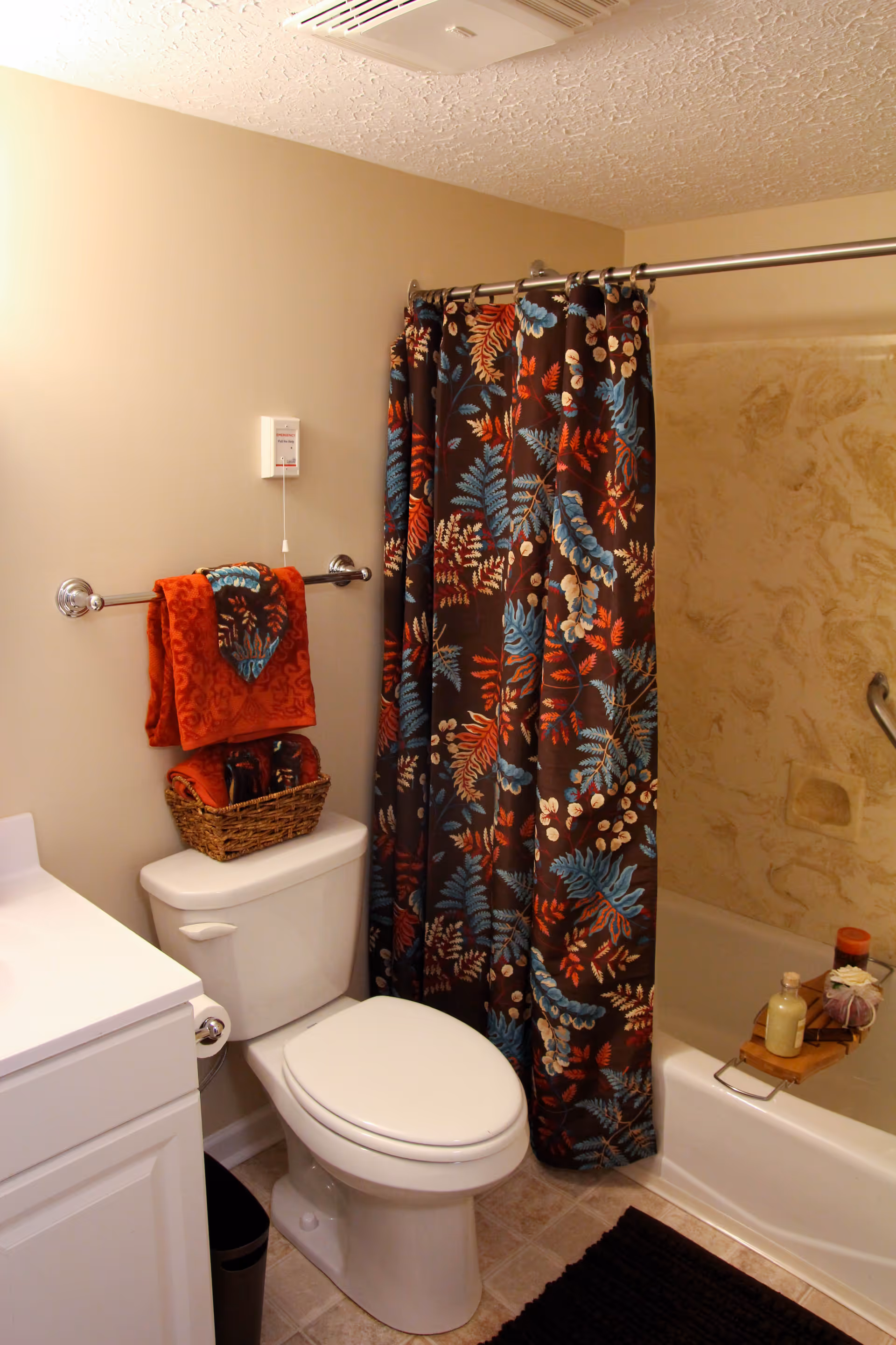 A bathroom with a white toilet, a white vanity with a sink, and a bathtub with a colorful floral shower curtain. There are red and patterned towels hanging on a towel rack above the toilet, and a small basket with towels on top of the toilet tank. The bathtub has a wooden tray holding bath items like soap and a bottle.
