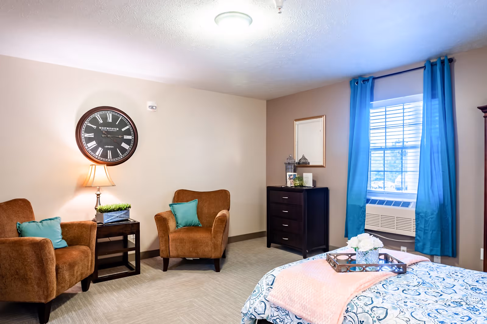 A cozy living area in a senior living facility with two brown armchairs each with a teal pillow, a small side table with a lamp and a plant, a large wall clock, a dresser with decorative items, a window with blue curtains, and a bed with a patterned bedspread and a pink throw blanket.