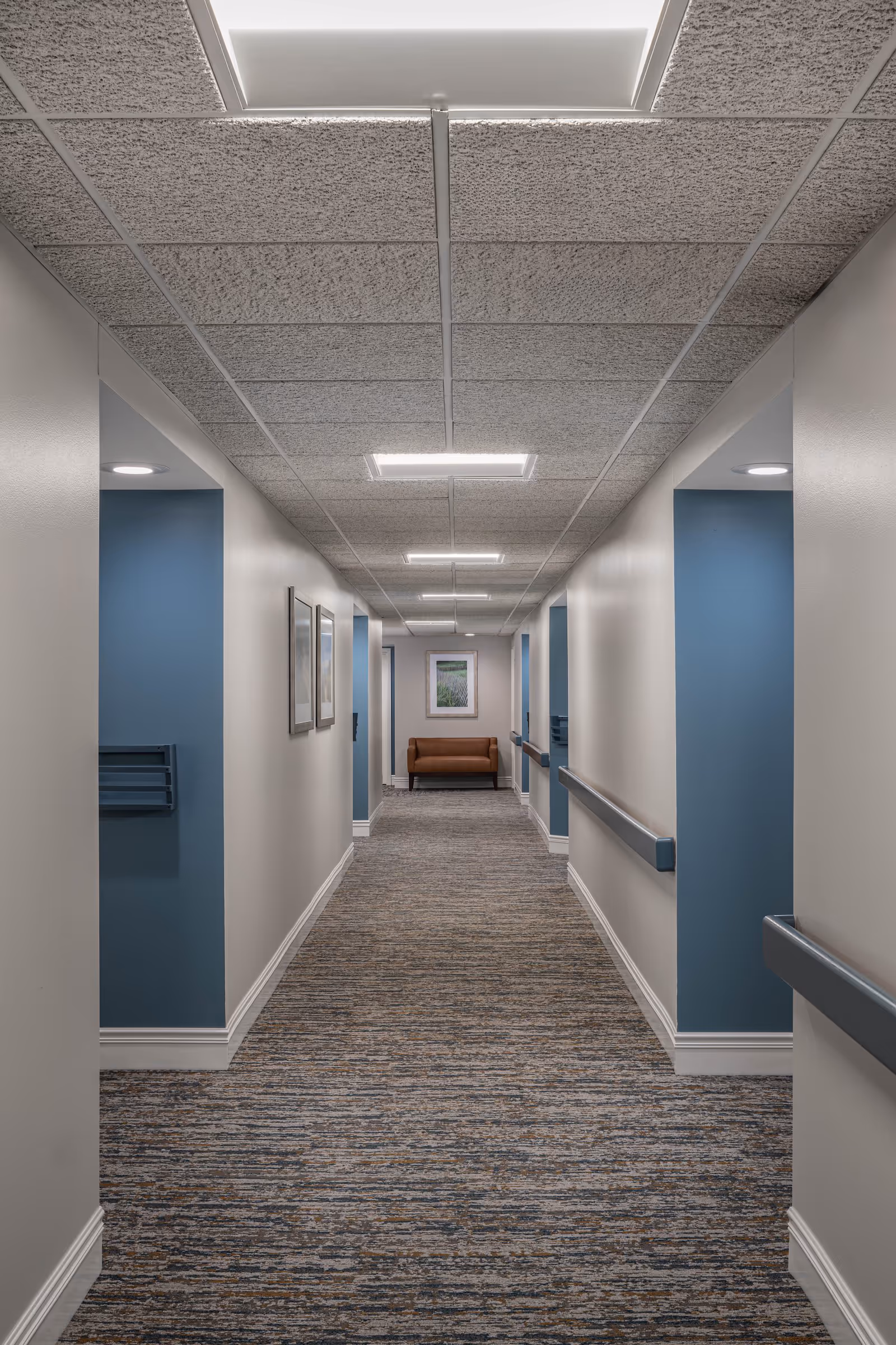 A long, well-lit hallway in a senior living facility with blue and white walls, carpeted floor, handrails on both sides, framed pictures on the left wall, and a brown bench at the far end beneath a framed picture.