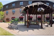 Outdoor courtyard area with a wooden gazebo, benches, and hanging flower baskets in front of a multi-story brick building with windows and red awnings.