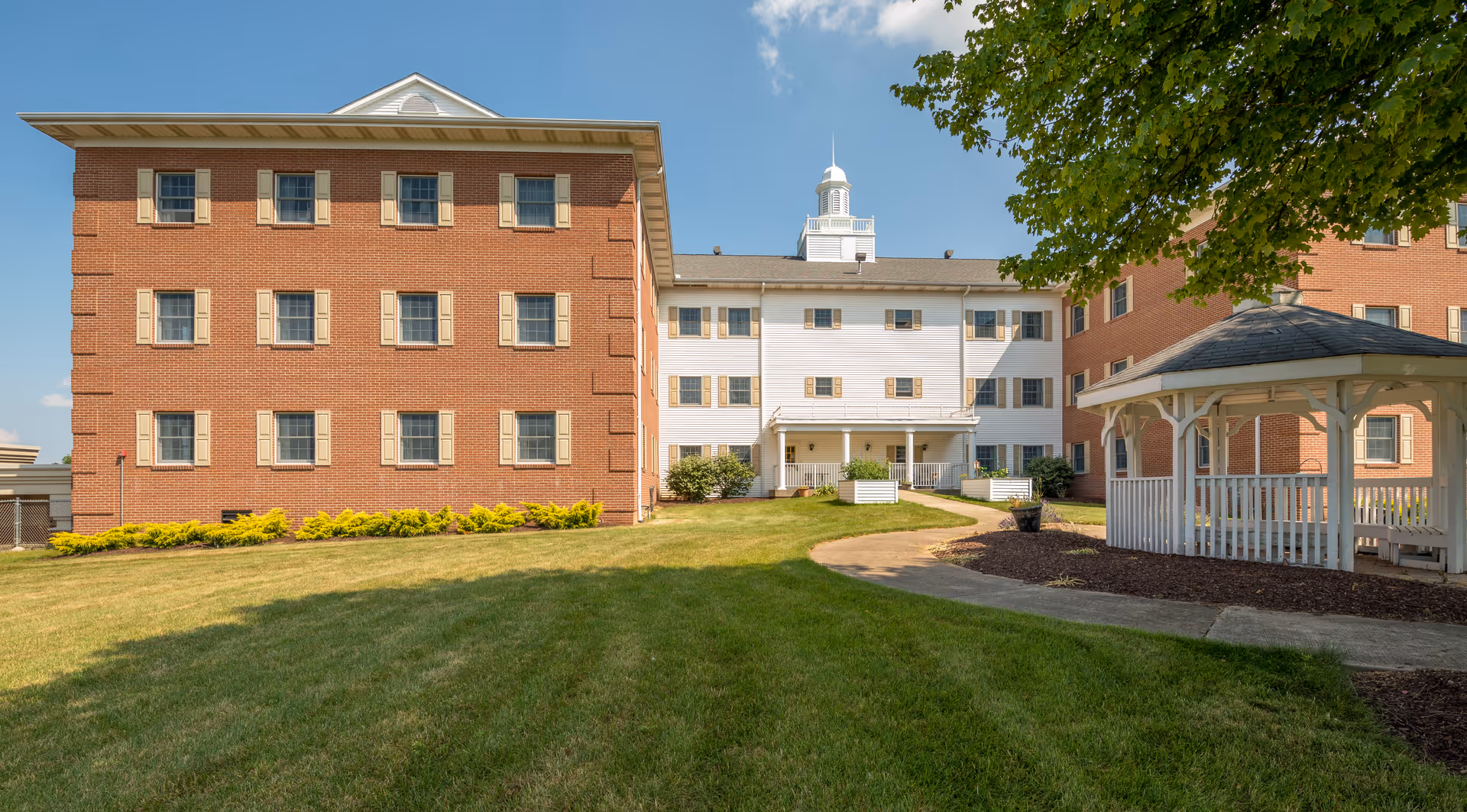 Exterior view of Shepherd of the Valley - Poland senior living facility showing a large brick and white building with multiple windows, a well-maintained grassy lawn, a paved walkway, and a white gazebo on the right side under a tree.