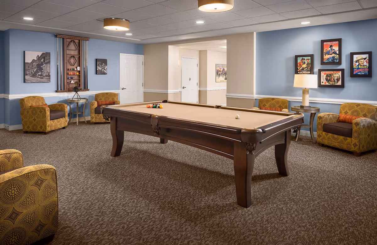 A cozy game room with a pool table in the center, surrounded by four patterned armchairs with orange cushions. The walls are painted light blue with white trim, decorated with framed vintage posters and black-and-white photographs. A lamp on a side table provides warm lighting, and a rack holding pool cues and balls is mounted on the wall.