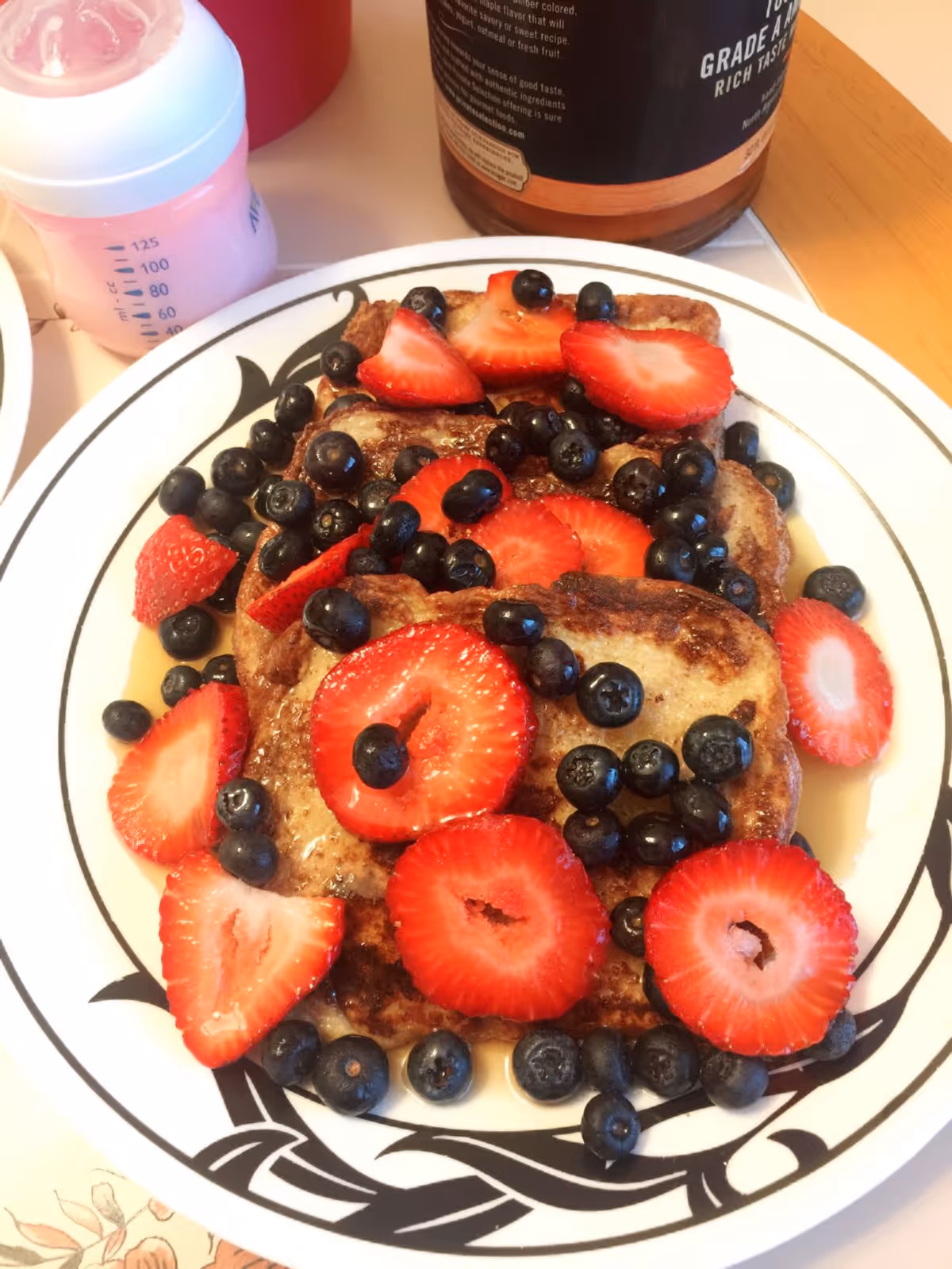 A plate of pancakes topped with fresh blueberries and sliced strawberries, with syrup drizzled over them. A pink baby bottle and a bottle of syrup are visible in the background.