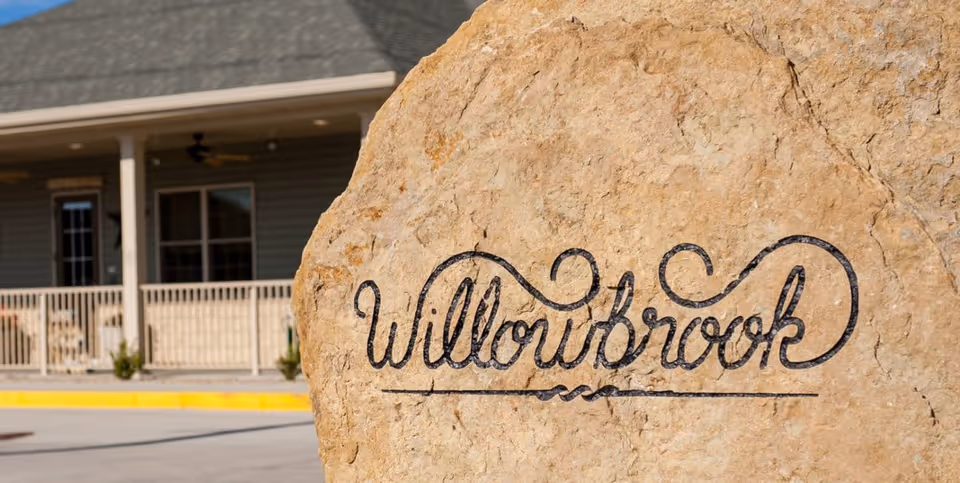 A large natural stone with the word 'Willowbrook' artistically engraved on it, placed outdoors in front of a building with a porch and railing.