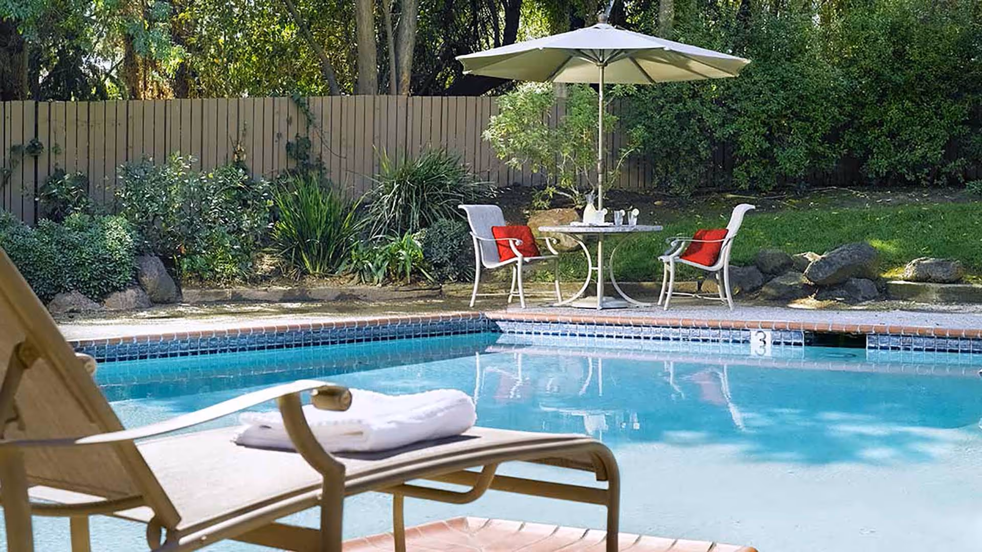 Outdoor swimming pool area with a lounge chair holding a folded white towel in the foreground. In the background, there is a small round table with two chairs, each with a red cushion, and a large white umbrella providing shade. The area is surrounded by greenery and a wooden fence.