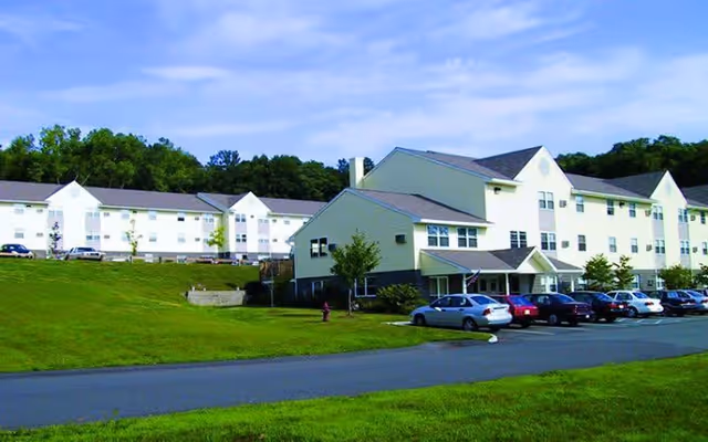 Exterior view of a large, multi-story senior living facility building with a parking lot in front and green grassy areas surrounding it under a partly cloudy sky.