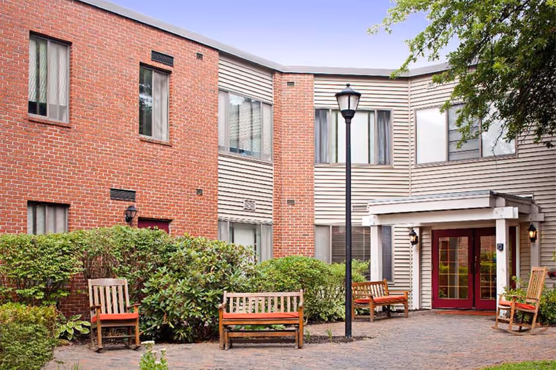 Outdoor courtyard area of Marshwood Center featuring a brick and siding building with multiple windows, wooden benches with red cushions, green bushes, a black lamppost, and a paved walkway under a clear blue sky.