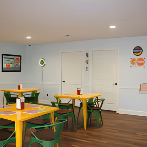 Interior view of a dining area with yellow tables and green chairs. The room has light blue walls, wooden flooring, two closed white doors, and some wall decorations including a framed picture and a circular artwork. The tables have placemats and condiment holders on them.