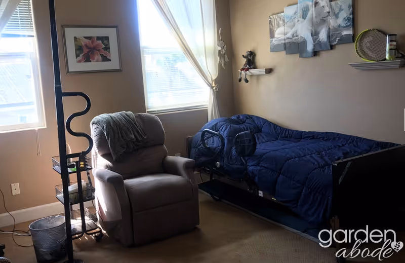 A cozy bedroom with a single bed covered in a blue comforter, a beige recliner chair with a gray blanket draped over it, two windows with white curtains letting in natural light, a framed flower picture on the wall, and decorative shelves holding small items. The room has beige walls and carpeted floor.