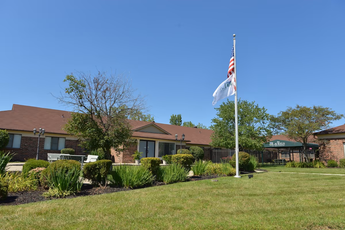 Exterior view of The Laurels of West Carrollton senior living facility with a well-maintained lawn, shrubs, trees, a flagpole with American and facility flags, and a brick building under a clear blue sky.