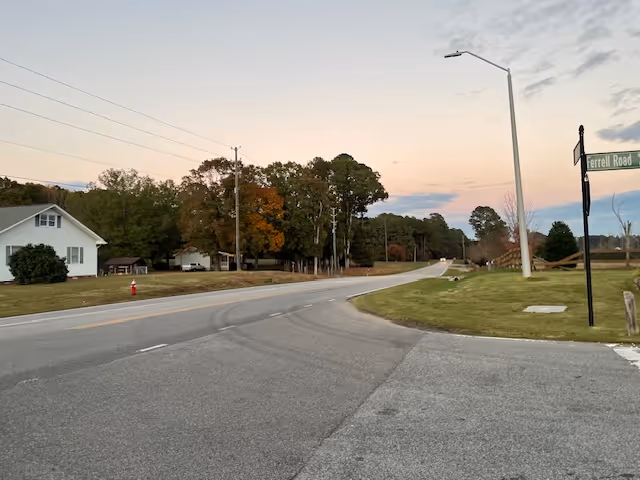 A quiet rural road at dusk with a street sign labeled Ferrell Road on the right side. There is a white house with a green bush in front on the left side, some trees with autumn foliage, a fire hydrant near the road, and a streetlight on the right. The sky is partly cloudy with soft pastel colors from the setting sun.