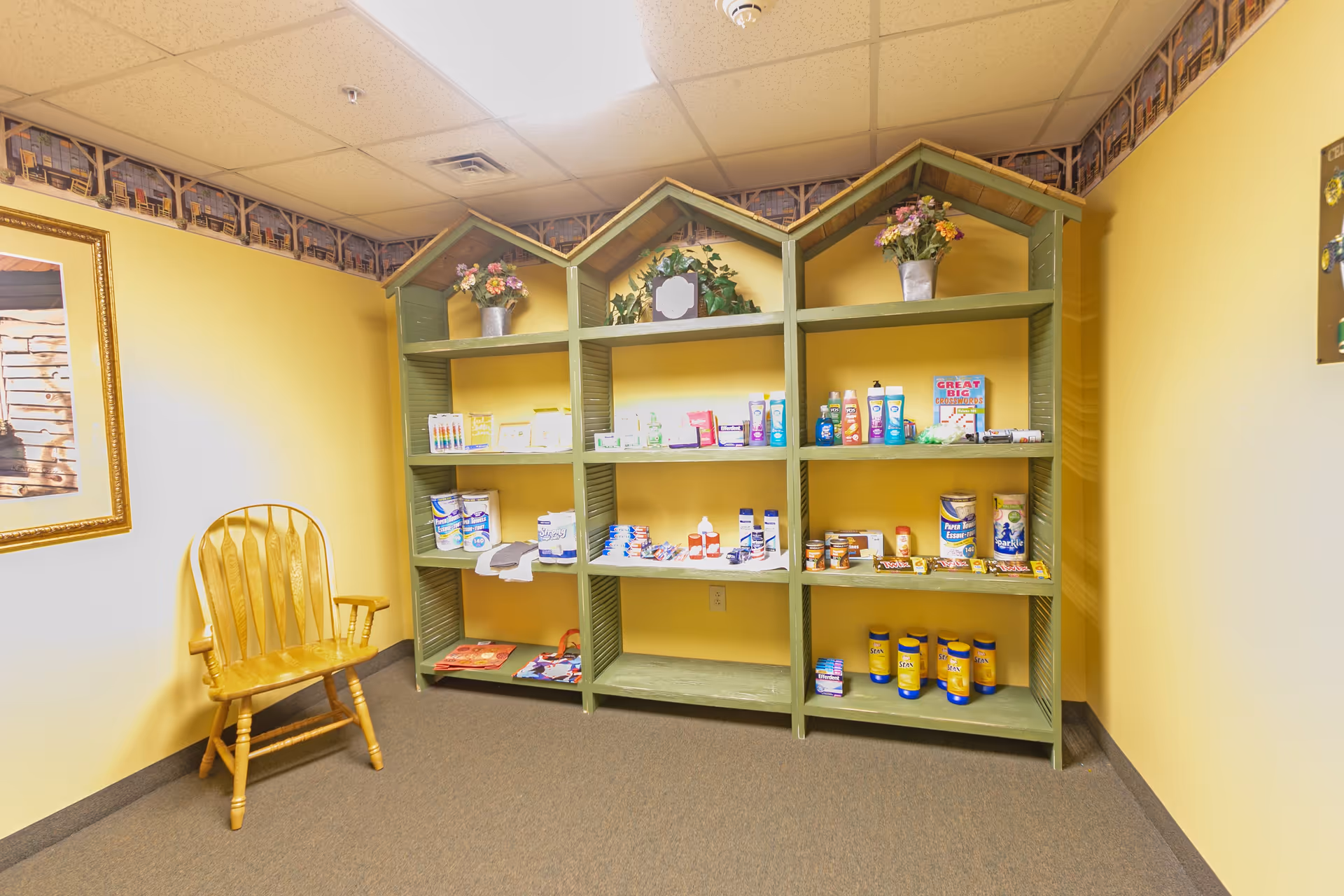 A small room with yellow walls and a brown carpeted floor featuring a green wooden shelving unit shaped like three small houses. The shelves hold various personal care items, toiletries, and snacks. To the left of the shelves is a wooden chair with armrests, and a framed picture hangs on the wall above it. The ceiling has white acoustic tiles with a fluorescent light fixture.