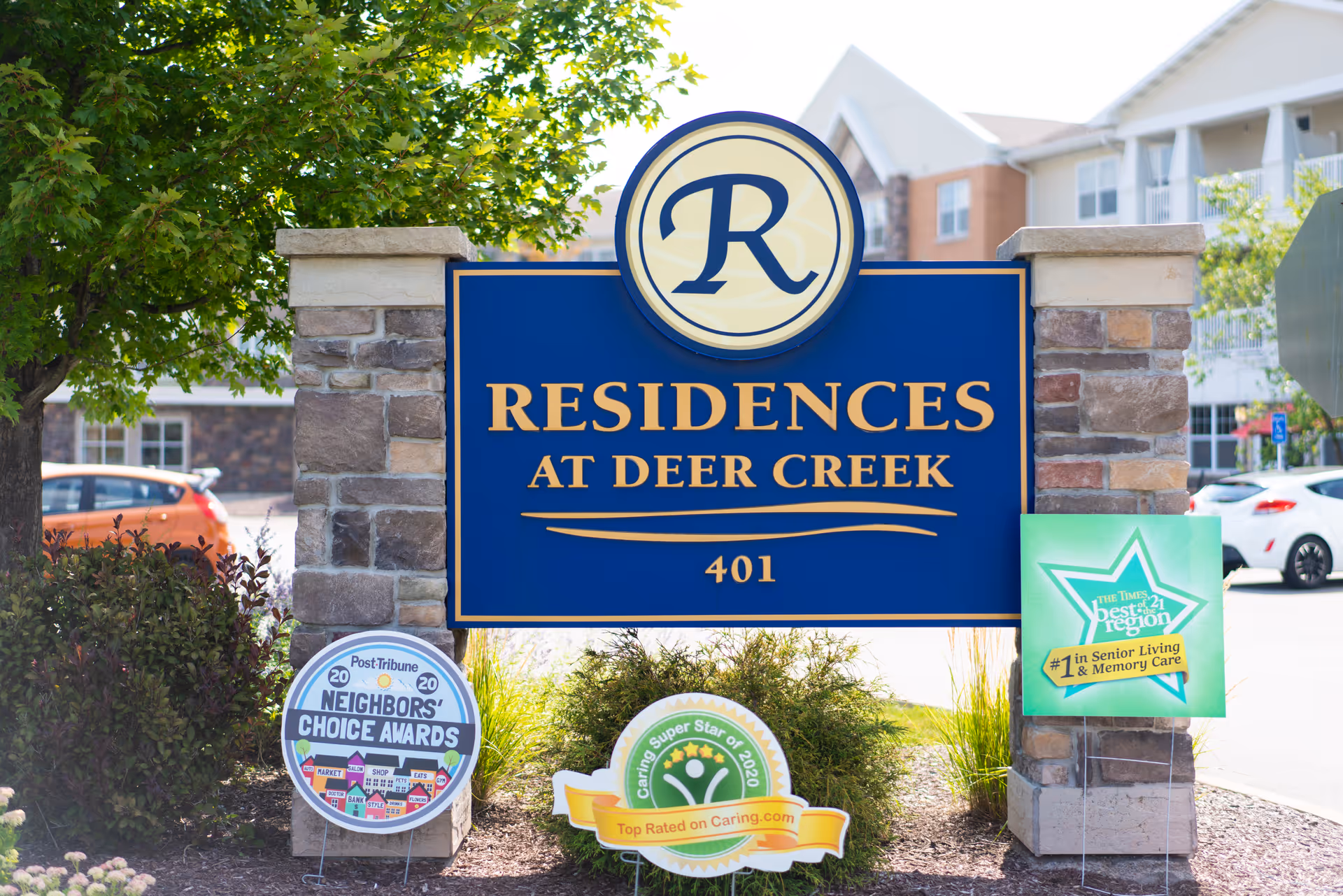 A large blue and gold sign for Residences At Deer Creek mounted between two stone pillars with greenery and awards displayed at the base. In the background, there are parked cars and a multi-story building.