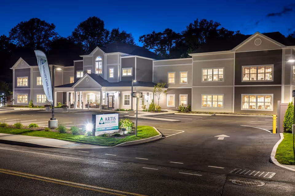 Exterior nighttime view of Artis Senior Living of Briarcliff Manor building with illuminated windows, a parking lot, and a sign in front displaying the facility's name and contact information.