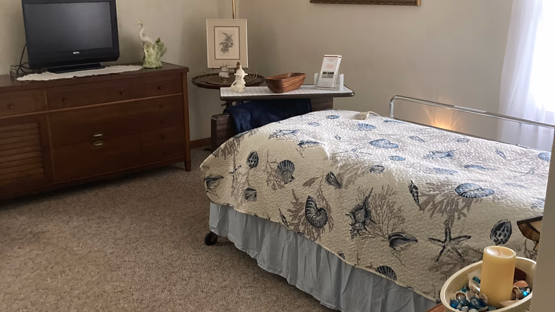 A cozy bedroom with a bed covered in a seashell-patterned quilt, a wooden dresser with a TV on top, a small round table with decorative items, and a window with sheer white curtains letting in natural light.