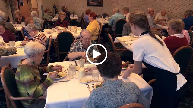 A dining room in a retirement community where elderly residents are seated at tables covered with white tablecloths, eating meals. A server in a white shirt and black apron is attending to one of the tables. The room is filled with people engaged in dining and conversation.