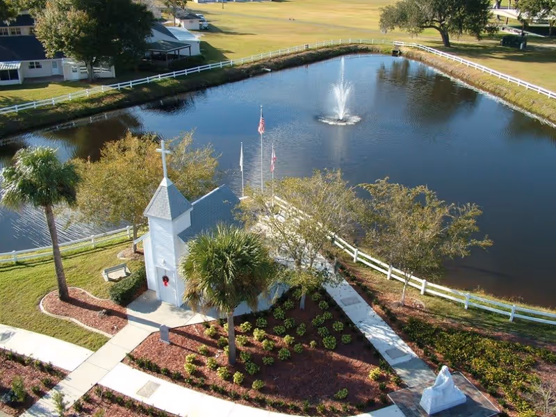 Aerial view of a small white chapel with a cross on top, surrounded by landscaped gardens and palm trees. The chapel is next to a pond with a water fountain in the center, bordered by a white fence. There are walkways leading to and around the chapel, and a statue is visible near the bottom right corner of the image.