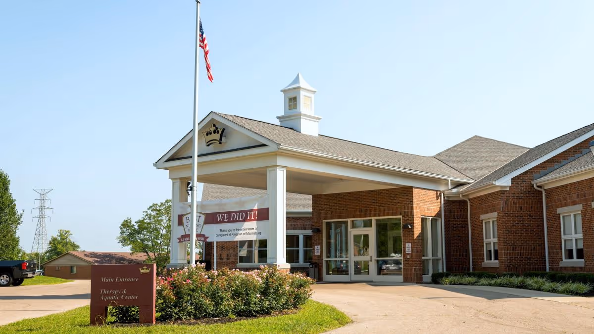 Exterior view of the main entrance of Kingston of Miamisburg, a brick building with a covered driveway, an American flag on a flagpole, and a sign indicating the Therapy & Aquatic Center. There is a banner hanging under the covered entrance that reads 'WE DID IT! Thank you to the entire team of caregivers at Kingston of Miamisburg.'