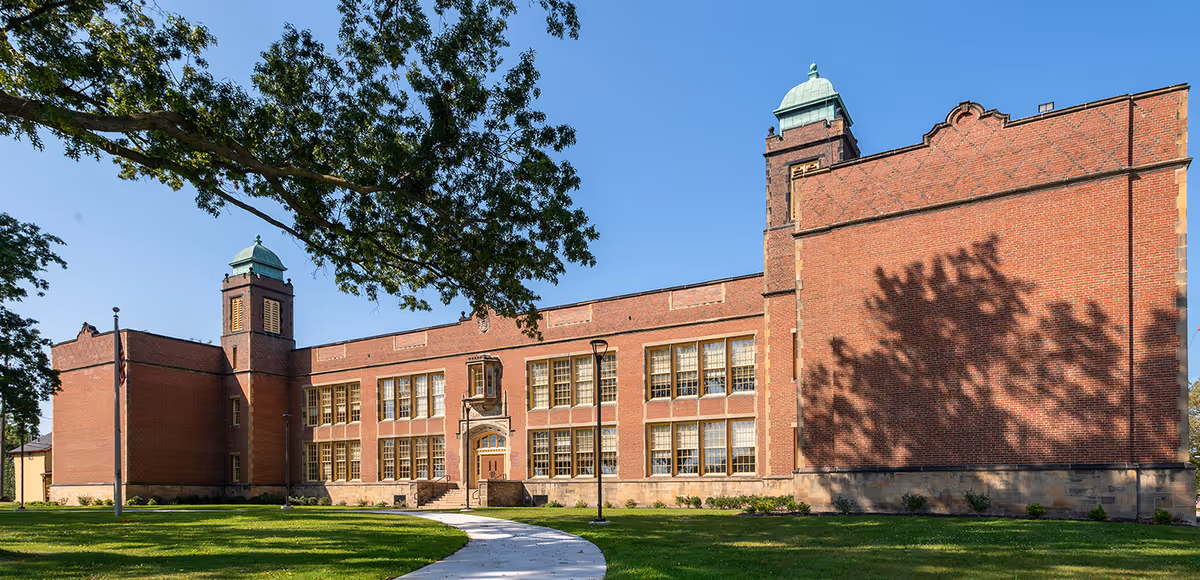 Exterior view of a large brick building with multiple windows and two towers topped with green domes. A curved concrete pathway leads to the main entrance, surrounded by green grass and trees under a clear blue sky.