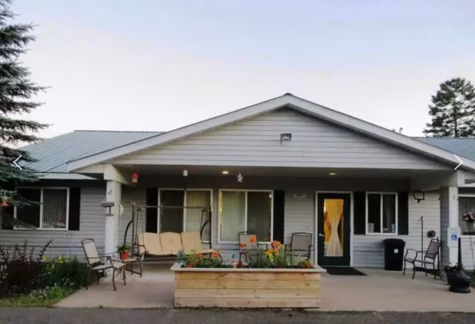 Exterior view of a single-story building with a covered porch area featuring outdoor seating including chairs and a swing. There is a raised wooden planter box with flowers in front of the porch. The building has light gray siding, a green door with a window, and several windows with white frames. Trees are visible in the background.