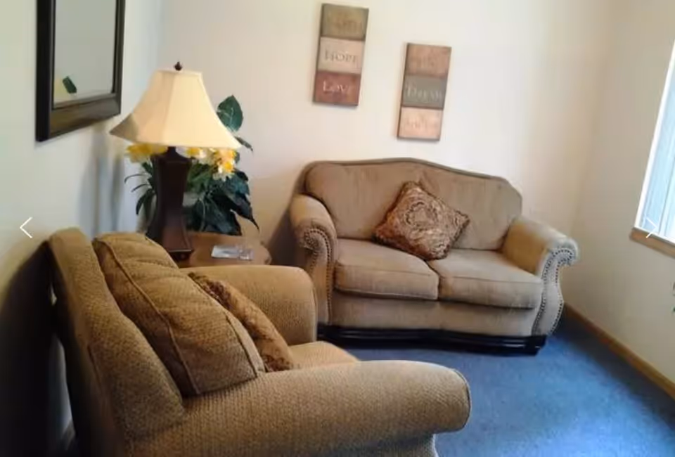 A cozy living room with two beige upholstered sofas, each with decorative pillows. There is a wooden side table between the sofas holding a lamp with a beige shade and a small plant. Two framed wall art pieces with inspirational words hang on the wall above the sofa. A window with vertical blinds allows natural light into the room.