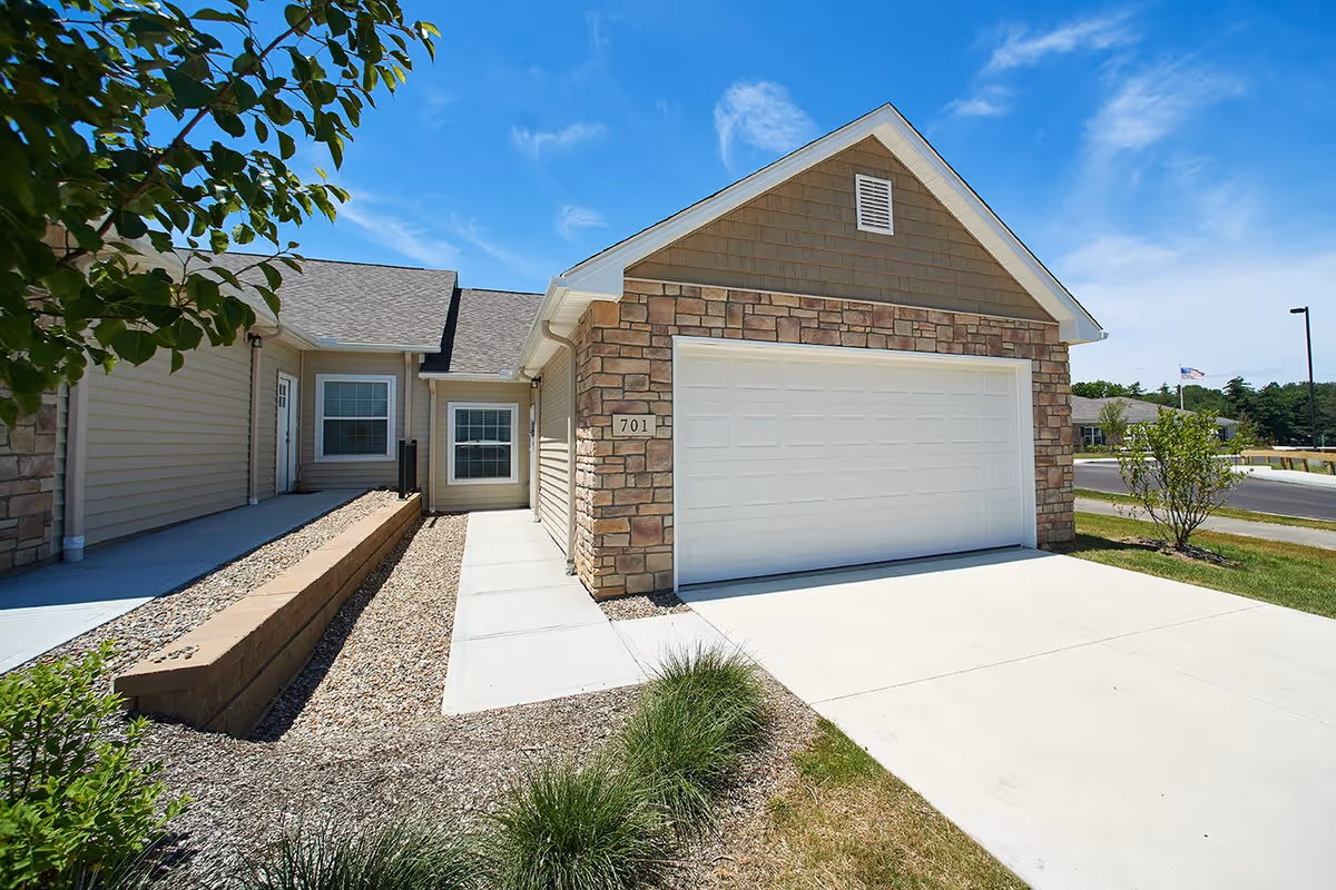 Exterior view of a single-story residential building with a stone and siding facade, a white garage door, and a concrete driveway under a clear blue sky.
