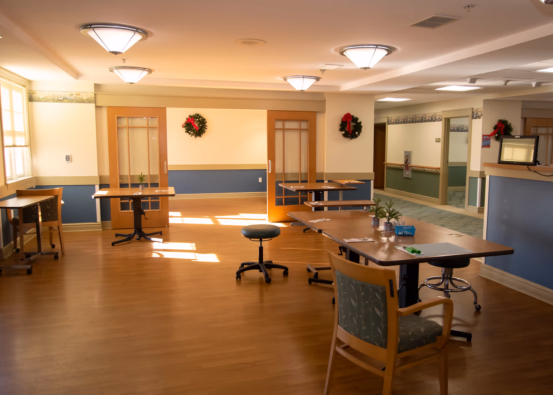 A well-lit room with wooden flooring and several tables and chairs arranged around the space. The walls are painted in a two-tone color scheme with blue on the lower half and cream on the upper half. There are two wreaths with red bows hanging on the walls, and a hallway with a mirror and handrails is visible to the right. The room has ceiling lights and large windows letting in natural light.