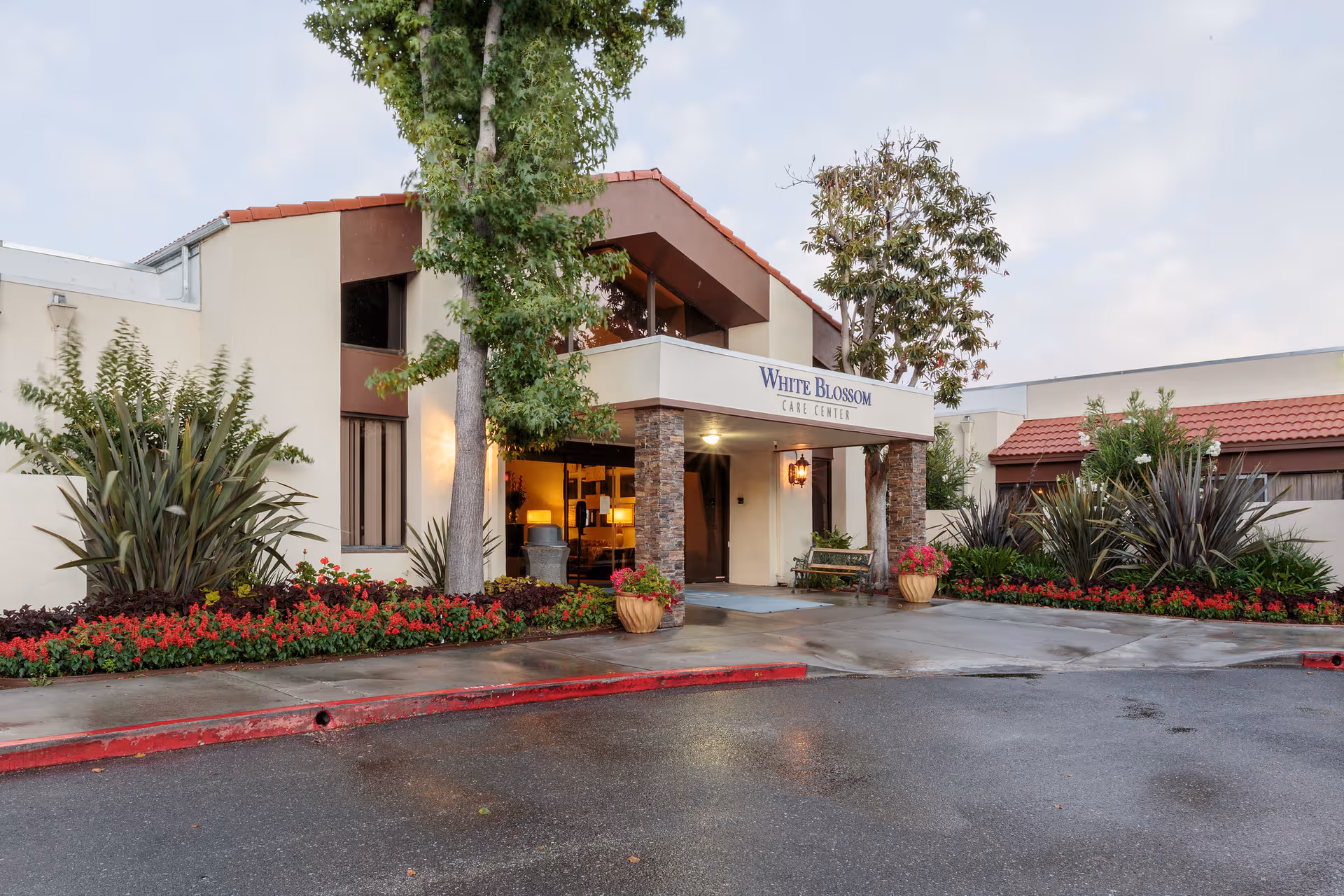 Exterior front entrance of White Blossom Care Center with a covered entryway supported by stone pillars, surrounded by landscaped plants and flowers, and a wet driveway in front.