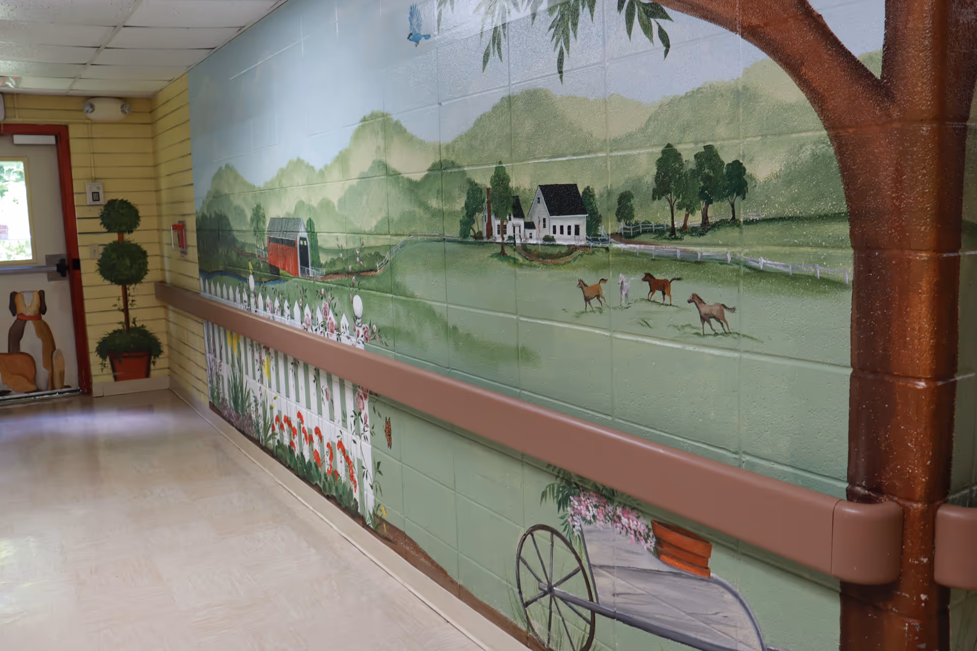 A hallway with a handrail on the right wall, which is decorated with a mural depicting a countryside scene including a white picket fence, flowers, horses, trees, a barn, and a house. On the left side near the door, there are two potted topiary plants and a painted dog figure on the door.