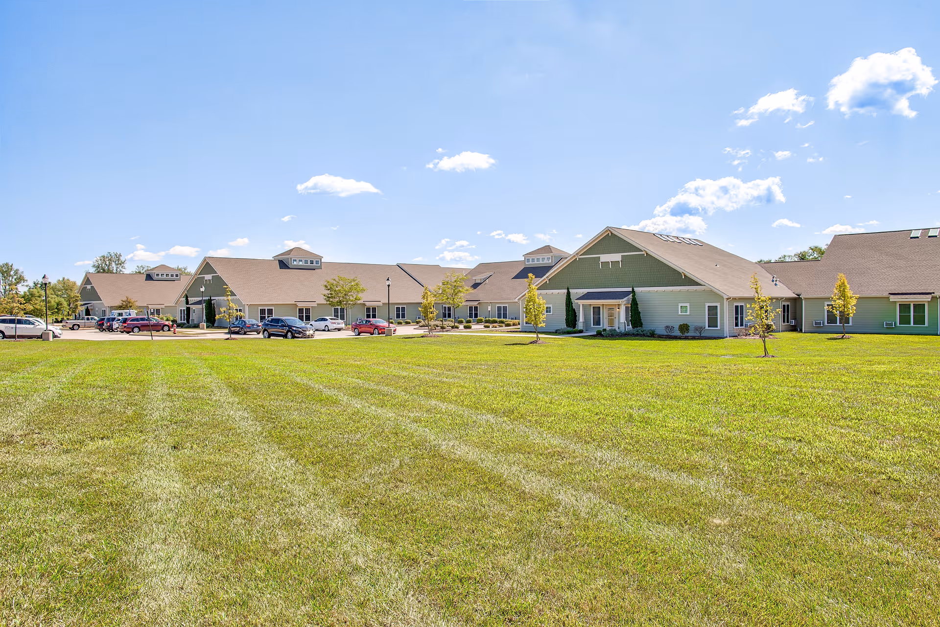 Wide view of a senior living facility building with a large green lawn in the foreground under a blue sky with scattered clouds. Several cars are parked near the building, which has a light green exterior and multiple peaked roofs.
