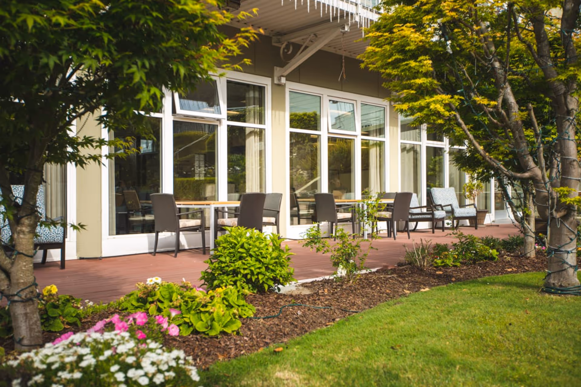 Outdoor patio area at a senior living facility with several tables and chairs arranged along a wooden deck. The patio is adjacent to large glass windows and doors, surrounded by green trees, shrubs, and colorful flowers in a landscaped garden.