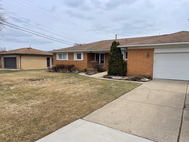 Single-story brick building with a sloped roof, a garage on the right side, a concrete driveway, and a lawn with some shrubs in front under a cloudy sky.