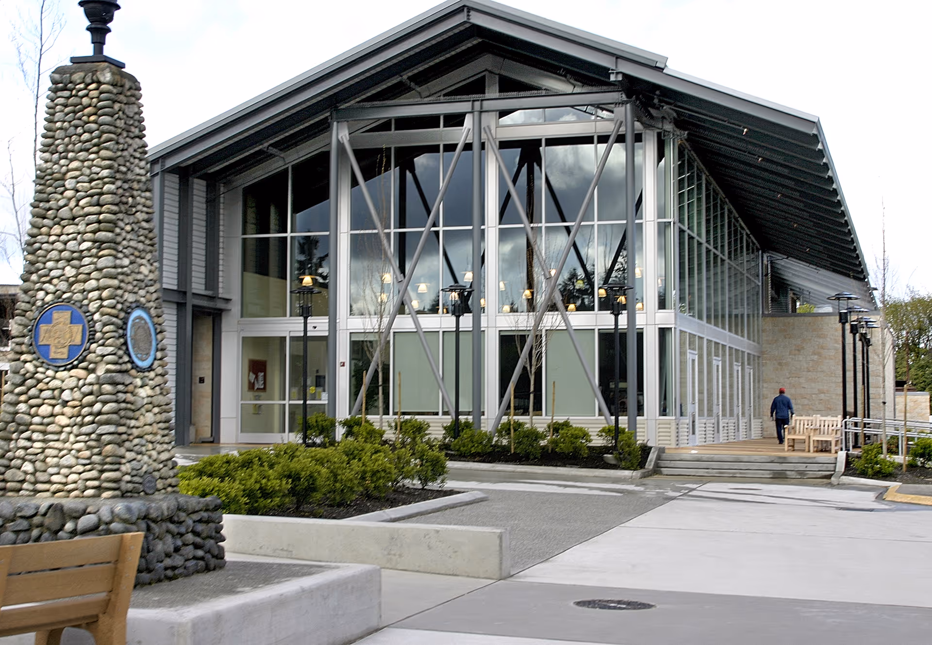 Exterior view of a modern building with large glass windows and a peaked roof. In front of the building is a stone monument with plaques, some greenery, benches, and a person walking near the entrance.